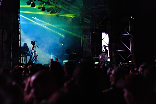 Menschenmenge bei einem Konzert mit Musikern auf der Bühne, die Instrumente spielen, unter Scheinwerferlicht, mit einem Banner und Metallrahmen im Hintergrund.