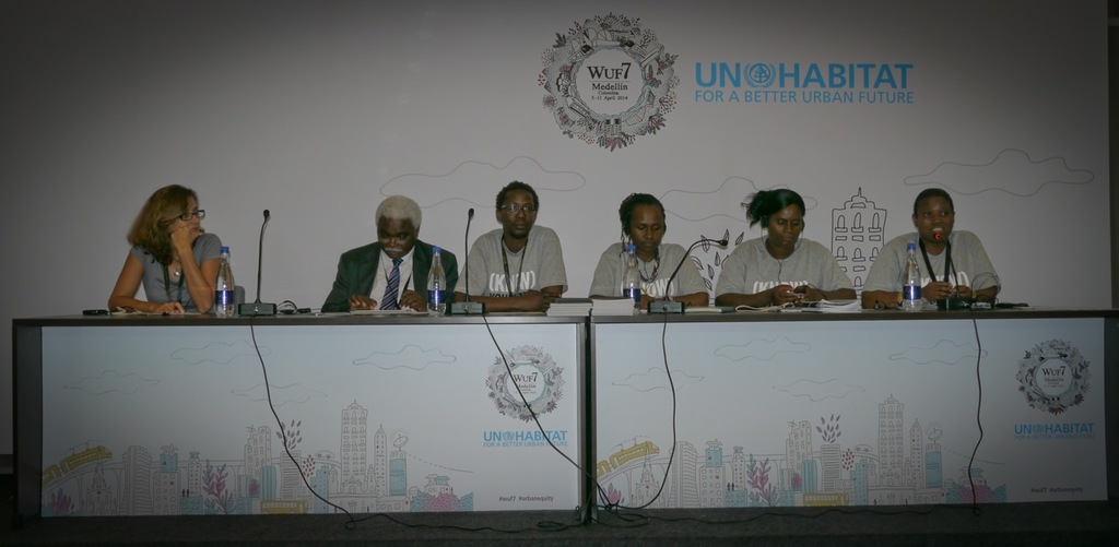 In einem Konferenzraum sitzen sechs Personen an zwei Tischen mit Büchern, Mikrofonen und Flaschen, eine Frau in einem grauen Kleid links, ein weißes Banner im Hintergrund und eine Flasche auf dem rechten Tisch.