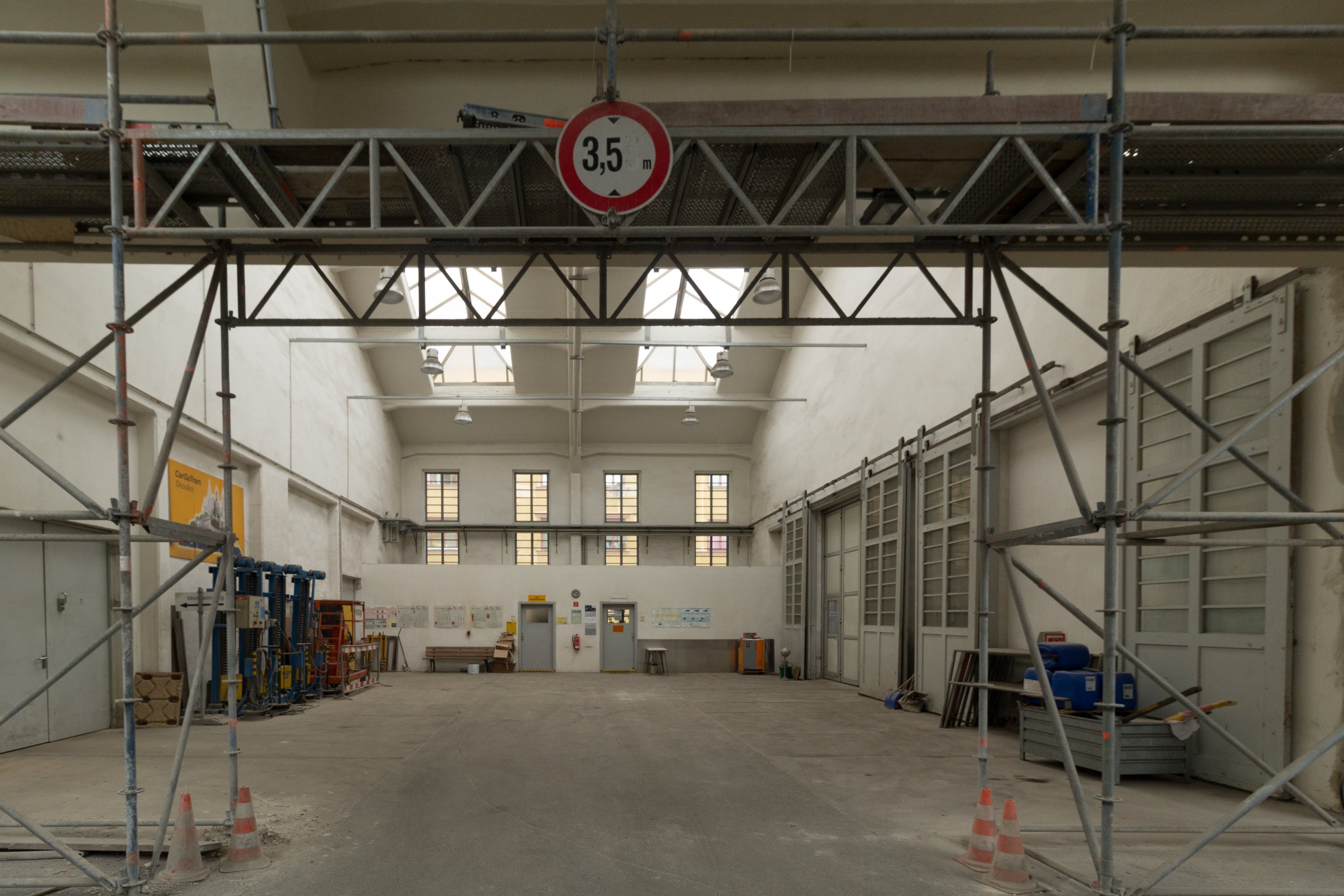 Großes Lagerhaus im Bau mit Gerüst, Verkehrskegeln, Metallstangen, einem Schild, Maschinen, einer Tür, Fenstern, Wänden, Deckenleuchten und einem Dach.