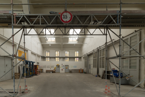 Großes Lagerhaus im Bau mit Gerüst, Verkehrskegeln, Metallstangen, einem Schild, Maschinen, einer Tür, Fenstern, Wänden, Deckenleuchten und einem Dach.