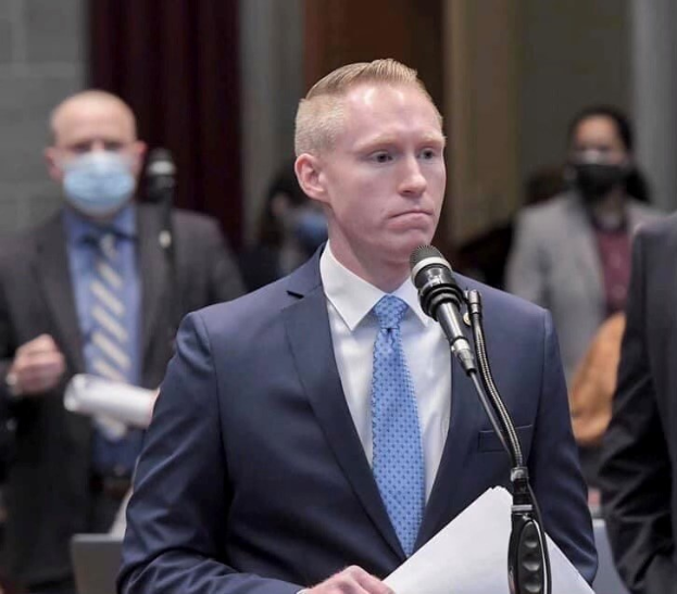 Ein Mann in Anzug und Krawatte spricht an einem Podium mit Papieren in der Hand, während andere in der Nähe stehen und ein Vorhang im Hintergrund zu sehen ist.