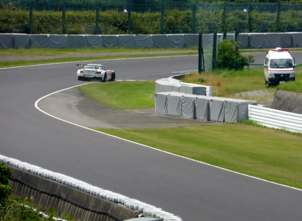 Ein Rennwagen fährt auf einer Rennstrecke, mit einem Polizeiwagen im Hintergrund, umgeben von Gras, Pflanzen, Bäumen und Zaun, mit einem Fahrzeug auf der rechten Seite und Text unten.