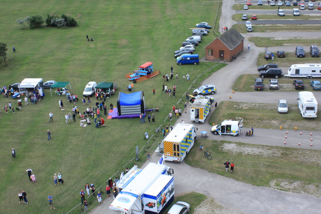 Eine Vogelperspektive eines Parkplatzes mit vielen Fahrzeugen, einigen Menschen in der Nähe von Zelten, einer zentralen Hütte und ein paar Bäumen auf der linken Seite.