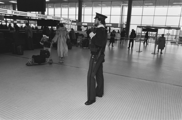 Polizeibeamter in Uniform mit Pistole in einem Flughafen-Terminal umgeben von Menschen und Gepäck, mit Gepäckwagen, Deckenleuchten, Glaswänden und Informationsanzeigen im Hintergrund.
