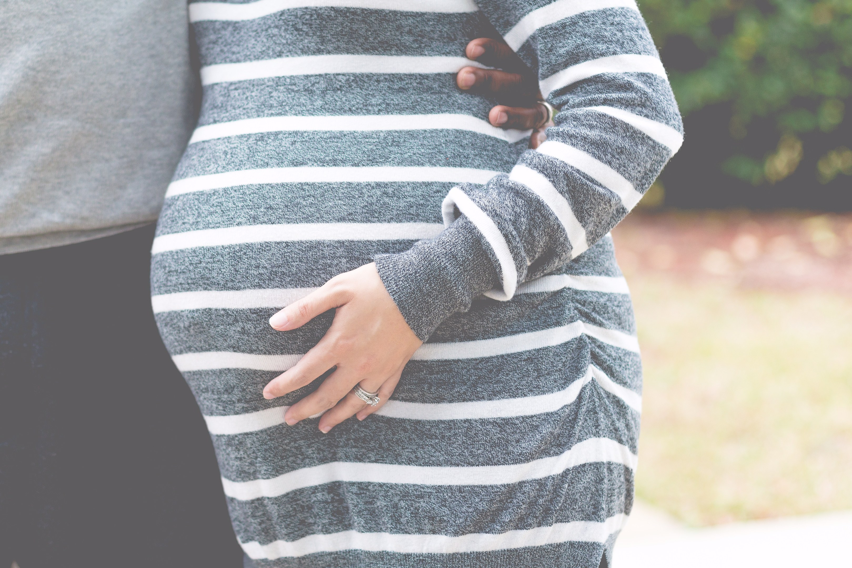 Schwangere Paare stehen zusammen mit den Händen auf ihrem Bauch, tragen Schwangerschaftskleidung, umgeben von Bäumen.