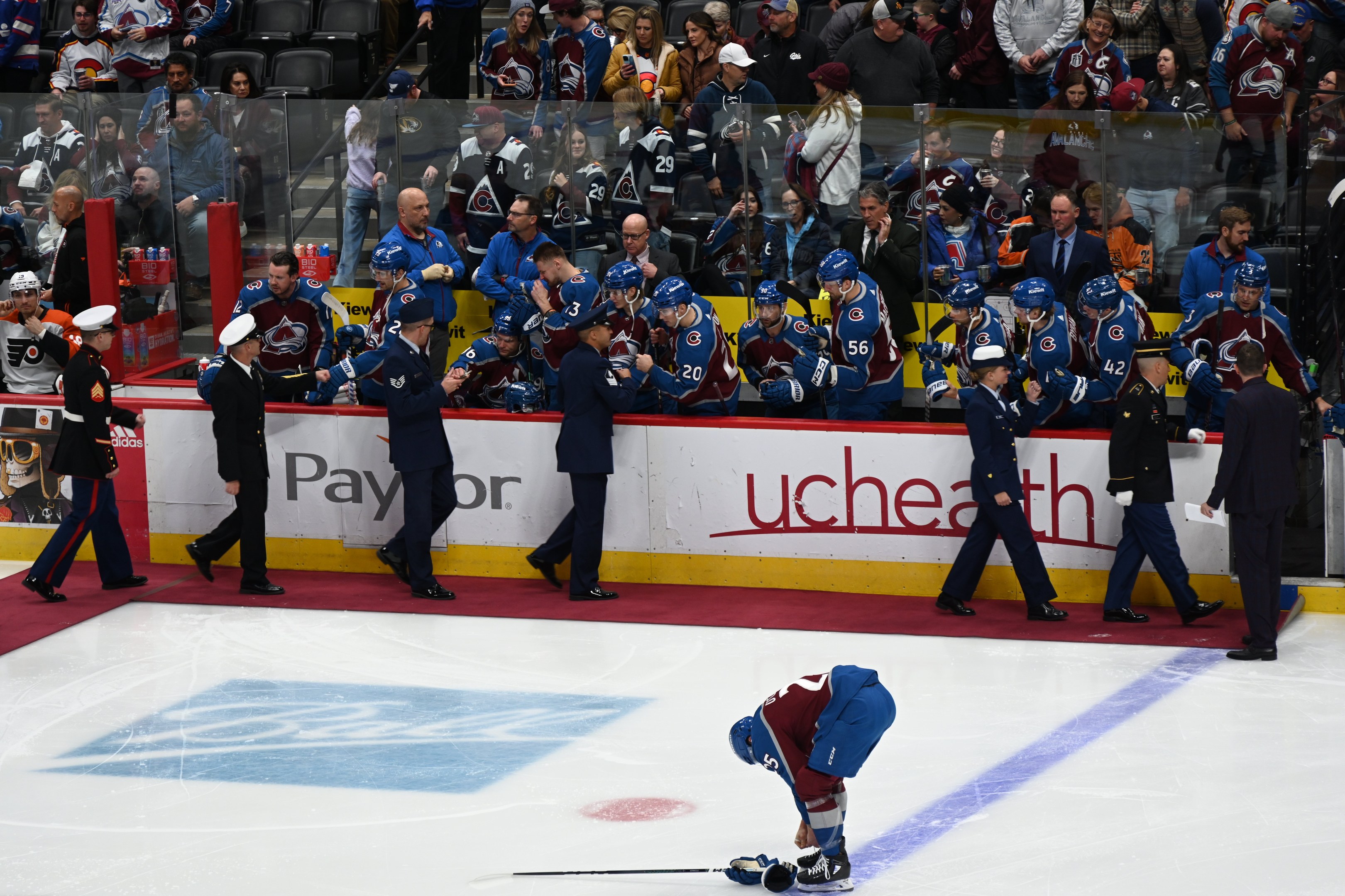 Eishockey-Spieler auf dem Eis umgeben von Teamkollegen und Gegnern, mit Zuschauern und Arena-Beschilderung im Hintergrund.