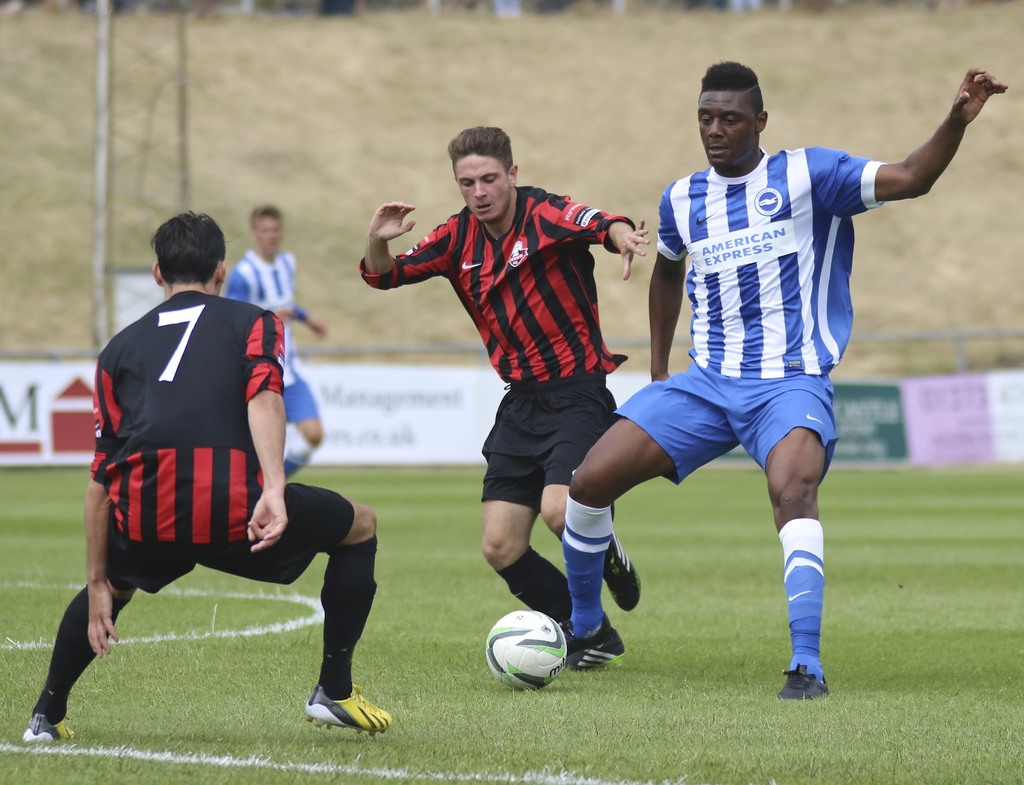 Vier Personen beim Fußballspielen auf einem grünen Feld, wobei eine Person in einem weißen und blauen Trikot rechts im Eck den Ball tritt und eine andere Person in der Mitte in rot-schwarzem Trikot steht.