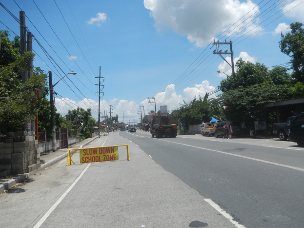 Eine Straße mit Fahrzeugen, ein "Slow Down School Zone"-Schild, Strommasten mit Drähten, Bäume, Laternenpfähle und ein bewölkter Himmel im Hintergrund.