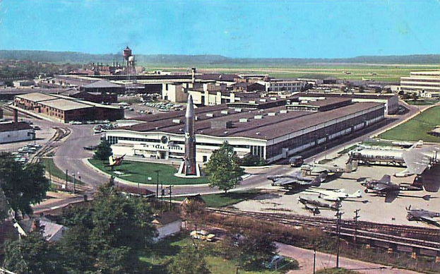 Alte Schwarz-Weiß-Flughafenszene mit geparkten Propellerflugzeugen, Terminalgebäuden, Straßen, Fahrzeugen, Strommasten, Grasflächen, Bahnschienen, fernen Hügeln und einem klaren Himmel; Bildtext unten.