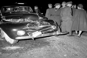 Ein Schwarz-Weiß-Bild eines auf der Straße liegenden, verbeulten Autos mit zersplitterten Fenstern, umgeben von einer Gruppe besorgter Menschen, die das Auto inspizieren.