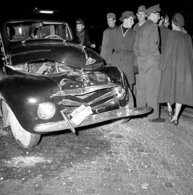 Ein Schwarz-Weiß-Bild eines auf der Straße liegenden, verbeulten Autos mit zersplitterten Fenstern, umgeben von einer Gruppe besorgter Menschen, die das Auto inspizieren.