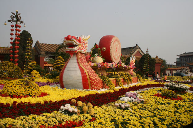 Ein chinesisches Neujahrsfest mit einer Drachenstatue, umgeben von bunten Blumen, Menschen, Gebäuden, Bäumen und einem klaren blauen Himmel im Hintergrund.
