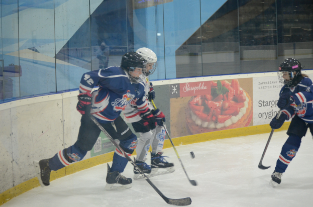 Gruppe junger Menschen, die Eis hockey auf einer Indoor-Eisbahn spielen, mit Helmen, Sportuniformen und Schlittschuhen, jeweils einen Hockey-Schläger haltend, mit einem Plakat im Hintergrund an einer Glaswand.