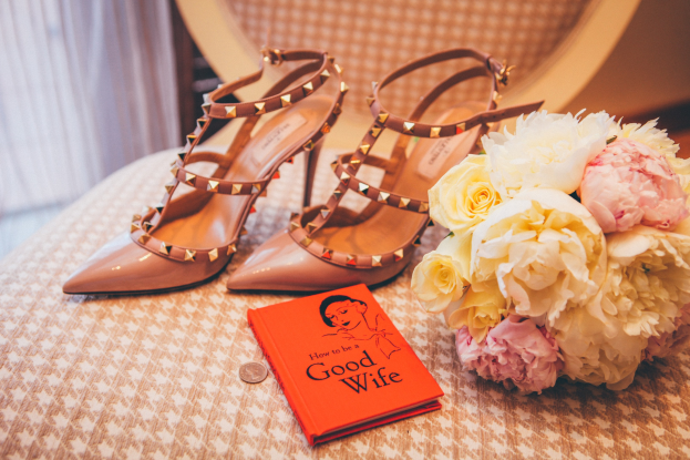 Ein Tisch zeigt ein Buch mit dem Titel "Wie man eine gute Ehefrau ist", einen Blumenstrauß und ein Paar Valentino-Schuhe, vor einem Vorhanghintergrund.