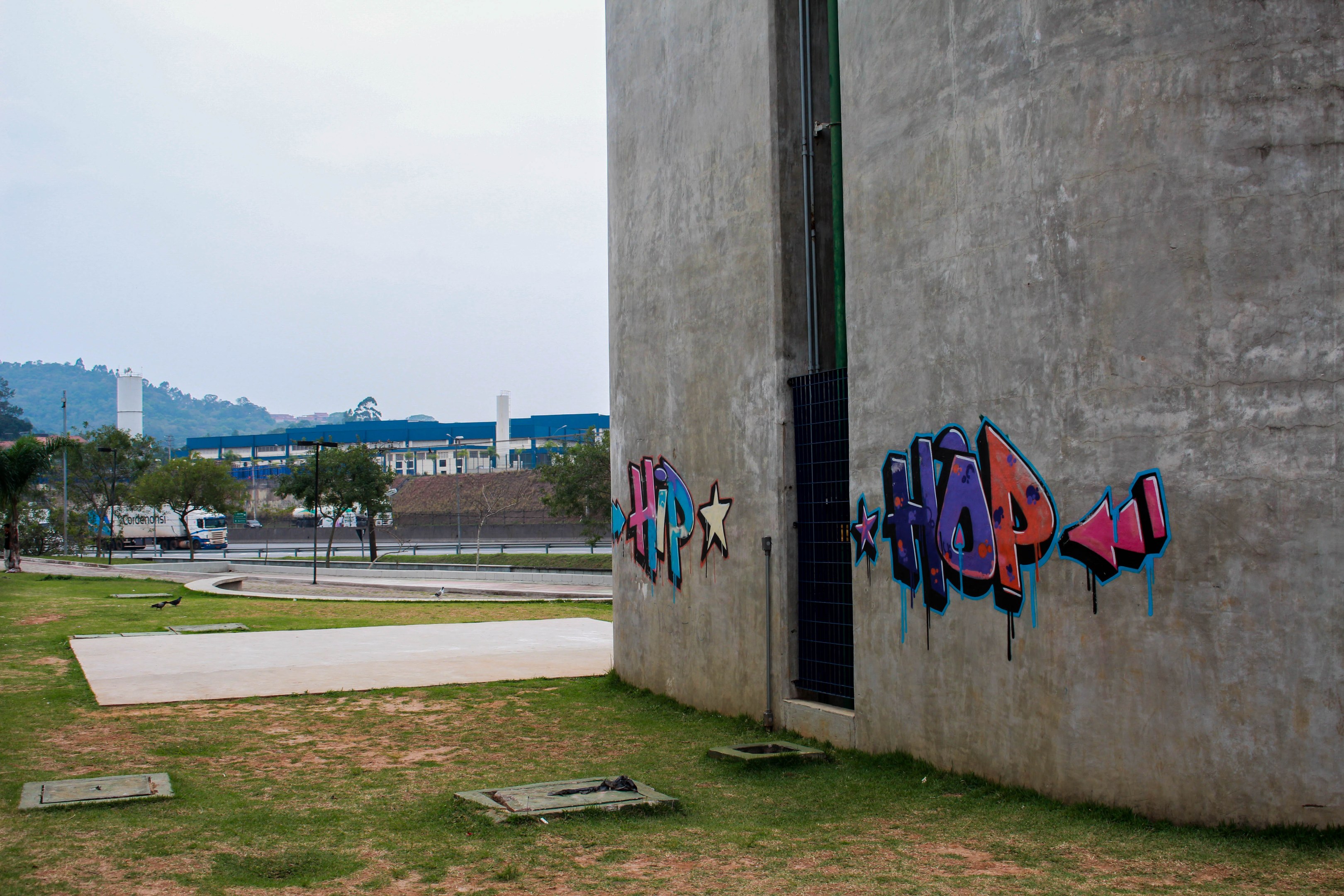 Betongebäude mit Graffiti, umgeben von Gras, Bäumen, Strommasten, Fahrzeugen, anderen Gebäuden, Hügeln und einem klaren blauen Himmel.