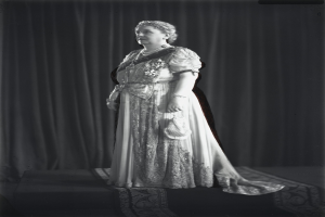 Schwarz-weiß-Foto der Queen Elizabeth II auf einer Bühne mit einem Vorhanghintergrund, die ein detailreiches königliches Kleid mit einem langen Rock trägt, gestyltes Haar in einem Dutt und einen sanften Ausdruck, der zur Seite schaut.