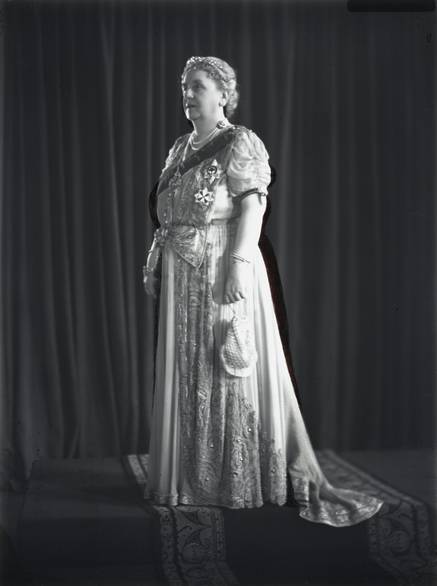 Schwarz-weiß-Foto der Queen Elizabeth II auf einer Bühne mit einem Vorhanghintergrund, die ein detailreiches königliches Kleid mit einem langen Rock trägt, gestyltes Haar in einem Dutt und einen sanften Ausdruck, der zur Seite schaut.