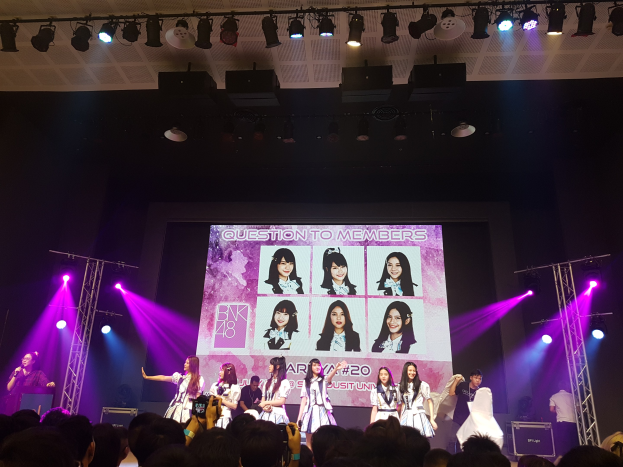 Eine Gruppe von Menschen, die auf einer hell erleuchteten Bühne performen, mit einer Menge darunter, einschließlich eines Sängers mit Mikrofon und eines Musikers, der ein Instrument spielt, während ein großer Bildschirm Bilder der K-Pop-Gruppe BK48 anzeigt.