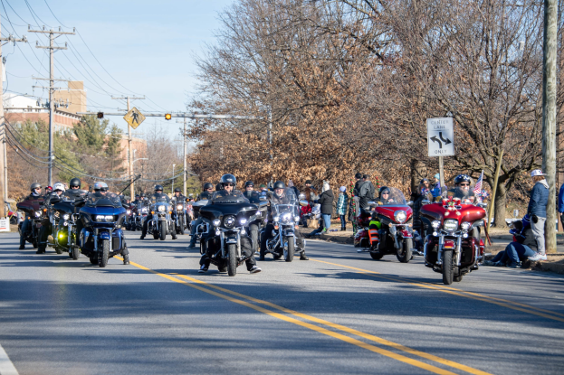 Eine Gruppe von Menschen auf Motorrädern fährt eine Straße entlang, gesäumt von Strommasten, Schildern, Bäumen und Gebäuden unter einem klaren blauen Himmel, wobei einige Fahrer Helme tragen.
