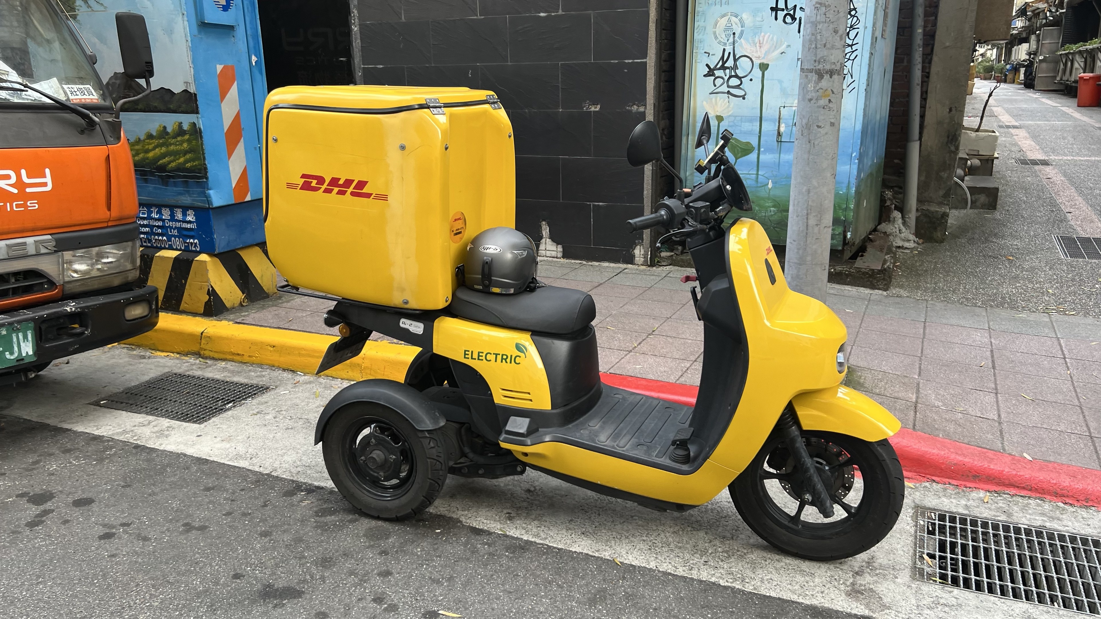 Ein gelber Elektro-Scooter mit Helm steht auf der Straße neben einem Lastwagen, mit Gebäuden und Menschen auf einem Gehweg im Hintergrund.