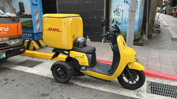 Ein gelber Elektro-Scooter mit Helm steht auf der Straße neben einem Lastwagen, mit Gebäuden und Menschen auf einem Gehweg im Hintergrund.