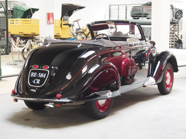 Schwarzes und rotes Auto in einer Garage mit anderen Fahrzeugen, Pfählen, Ketten und Gegenständen geparkt; Feuerlöscher an der Wand im Hintergrund.