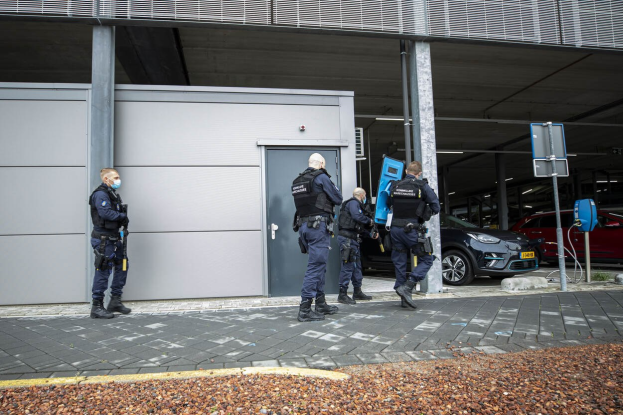 Eine Gruppe von Polizisten steht in einem Parkplatz vor einem Gebäude mit einer Tür, umgeben von Fahrzeugen, Pfählen, Platten und verstreuten Steinen.