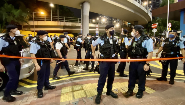 Polizisten in Uniform und Masken vor einer Menge, mit Fahrzeugen, Gebäuden, Bäumen und einer Brücke im Hintergrund.
