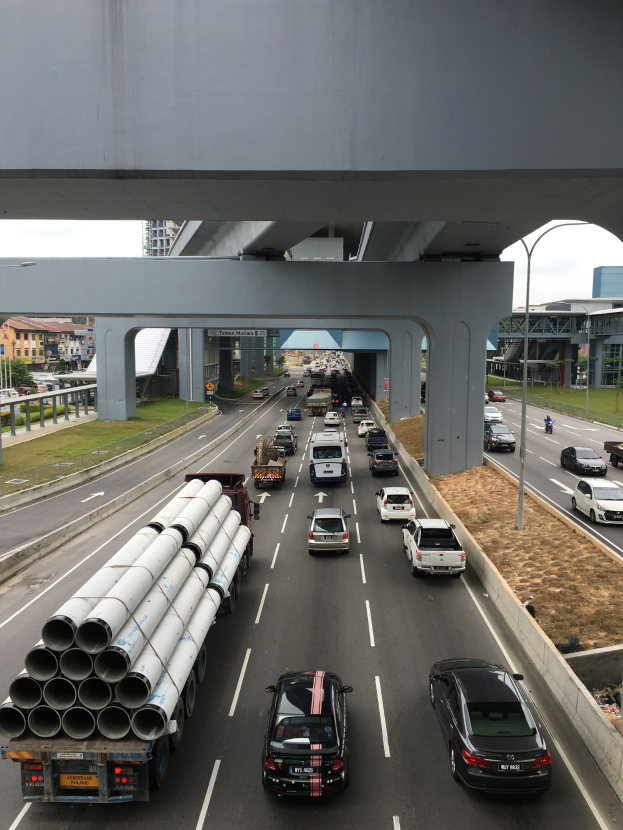Eine verkehrsreiche Autobahn mit Schwerlastverkehr unter einer Brücke, umgeben von Gras, Bäumen, Gebäuden und Strommasten unter einem klaren Himmel.