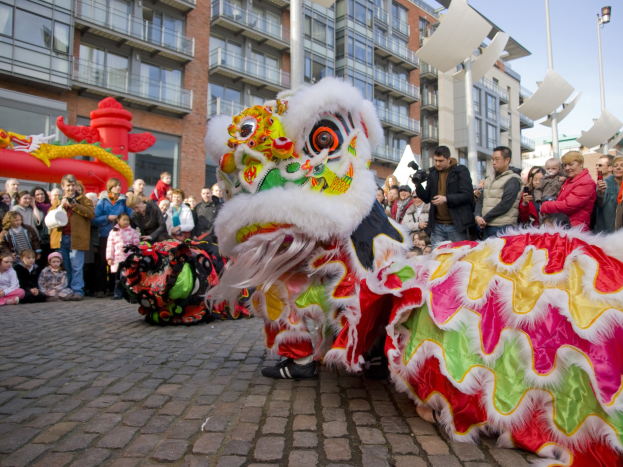 Ein lebendiges chinesisches Neujahrsfest in Amsterdam mit einer Löwen-Tanzvorstellung und einer Menge Schaulustiger, einige halten Kameras, vor einem Hintergrund aus Gebäuden, Laternenmasten und einem klaren blauen Himmel.