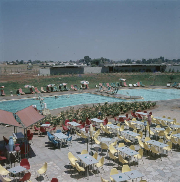 Ein Freibad mit Tischen, Stühlen und Sonnenschirmen umgeben von Blumenpflanzen, Pfählen, einem Zaun, einer Wand, einer Baumgruppe, einigen Gebäuden und einem bewölkten Himmel.