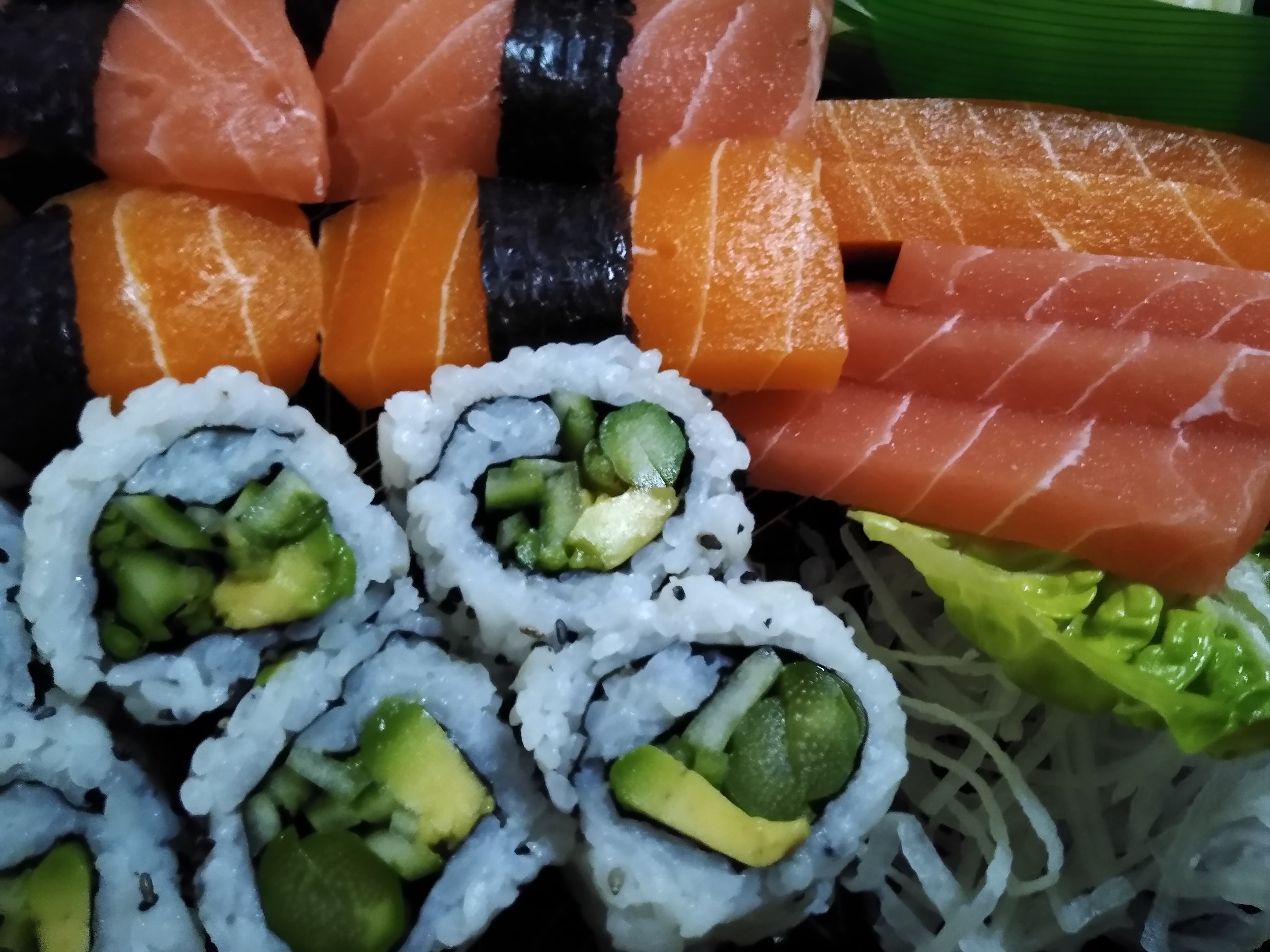 Ein schwarzer Teller mit verschiedenen Sushi-Rollen, darunter Lachs, Avocado und Gurke, garniert mit ein paar Blättern.