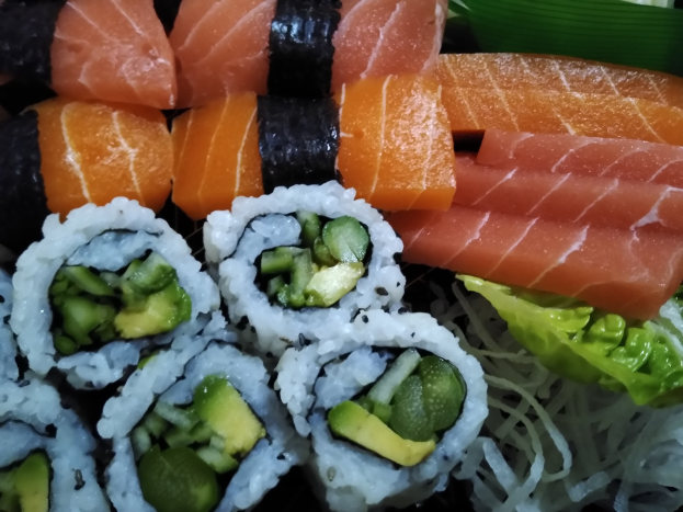 Ein schwarzer Teller mit verschiedenen Sushi-Rollen, darunter Lachs, Avocado und Gurke, garniert mit ein paar Blättern.