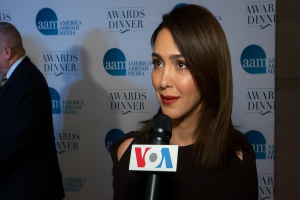 Frau in einem schwarzen Kleid mit einem Mikrofon, identifiziert als Emmy Rossum, mit einem Mann links von ihr und einer Tafel mit der Aufschrift "Aam Awards Dinner" im Hintergrund.