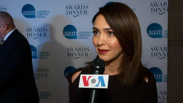 Frau in einem schwarzen Kleid mit einem Mikrofon, identifiziert als Emmy Rossum, mit einem Mann links von ihr und einer Tafel mit der Aufschrift "Aam Awards Dinner" im Hintergrund.