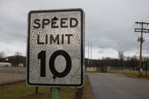 Ein Geschwindigkeitsbegrenzungsschild am Rande einer Stra├če, umgeben von Gras, Strommasten mit Kabeln, einem Zaun, H├Ąusern, B├Ąumen und einem bew├Âlktem Himmel im Hintergrund.
