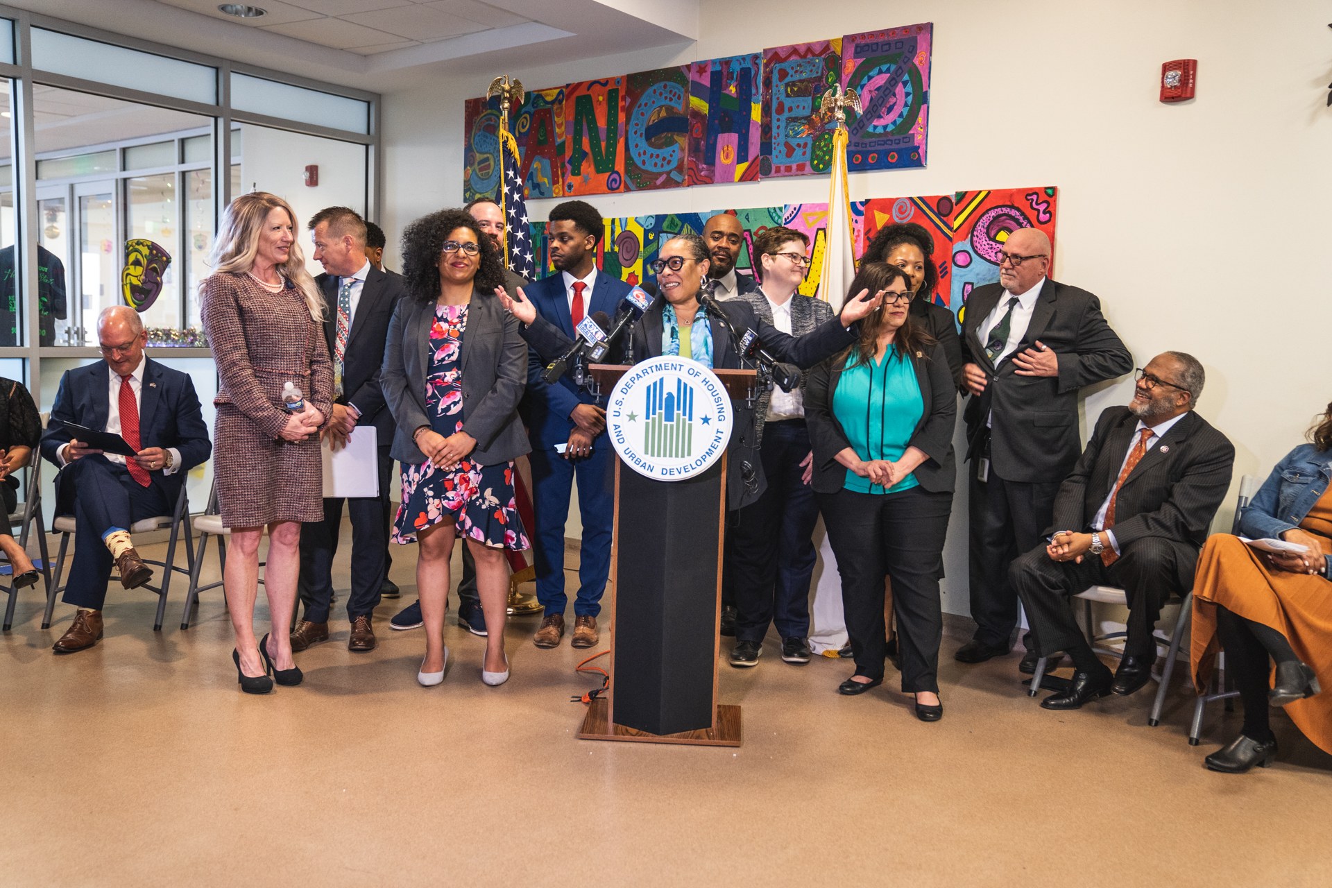 Gruppe von Menschen vor einem Podium mit Mikrofon und einer Tafel mit der Aufschrift "Sancho Stadtrat tritt dem Stadtrat bei", mit sitzenden und stehenden Teilnehmern, Fahnen, Feuerlöscher und Deckenbeleuchtung im Hintergrund.