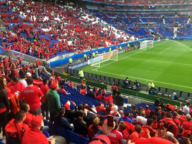 Ein vollbesetztes Stadion mit Sitzplätzen und Stehplätzen, Fahnen, einem Tor und Deckenleuchten, während die Menge ungeduldig auf den Beginn eines Fußballspiels wartet.