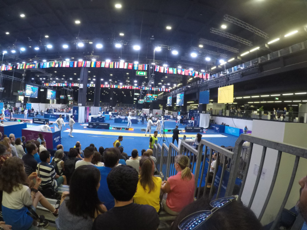Große Menschenmenge sitzend und stehend in einer Indoor-Sportarena mit Bildschirmen, Fahnen und Deckenbeleuchtung.