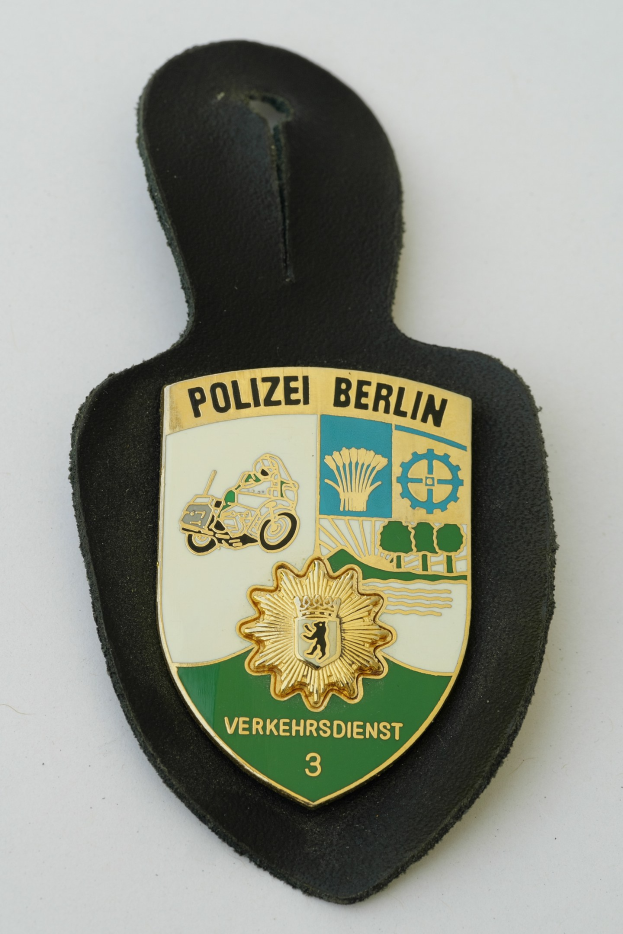 Ein schildförmiges Polize abadge mit "Polizei Berlin" in schwarzer Schrift, das einen blauen Hintergrund, einen weißen Stern und einen weißen Rand hat und auf einer Oberfläche liegt.