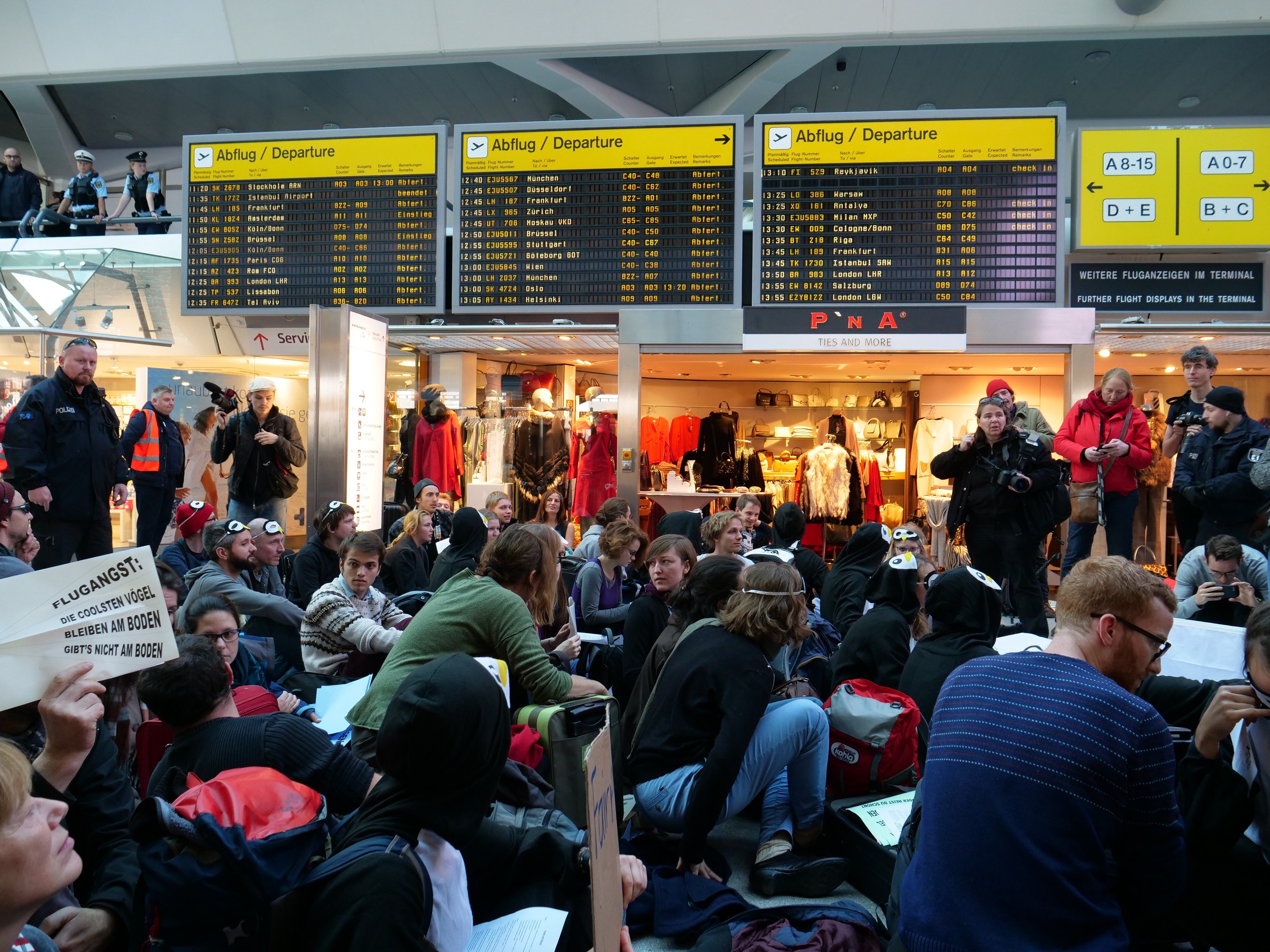 Große Gruppe von Menschen in einem Flughafen-Terminal, einige sitzen mit Taschen und Papieren, andere stehen, mit Informationsschildern, Schaufensterpuppen in Kleidern und Deckenbeleuchtung im Hintergrund, was auf eine Protestaktion hindeutet.