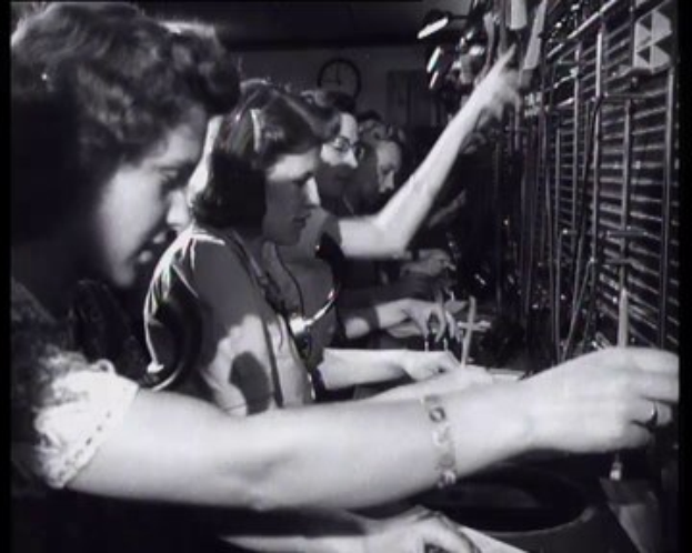 Schwarz-weißes Bild von Frauen, die vor einem Computer und einer Maschine stehen, mit einer Wanduhr im Hintergrund.