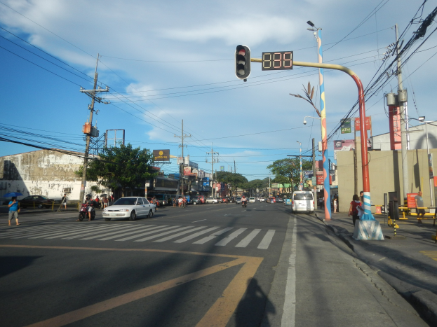 Eine Stadtstraße an einer Kreuzung mit einer Ampel, Fahrzeuge, Fußgänger, Strommasten, Laternen, Bäume, Gebäude und Schilder unter einem bewölkten Himmel.