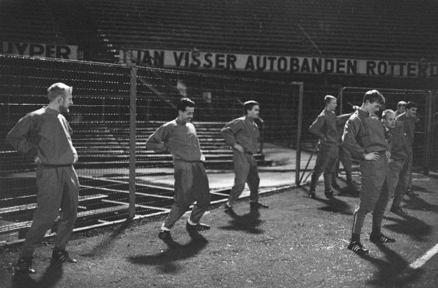 Schwarzes Bild einer Gruppe von uniformierten Männern, die nachts eine Straße entlanggehen, mit Stadionbeleuchtung im Hintergrund.