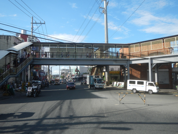 Stadtstraße mit Fahrzeugen, eine Fußgängerbrücke mit Menschen, Strommasten und Drähte, Gebäude und ein bewölkter Himmel.