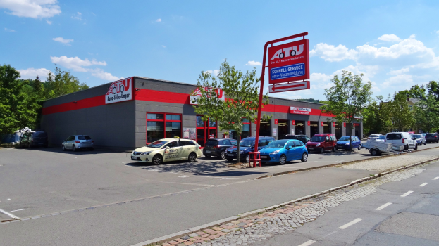 Ein Autohaus mit mehreren geparkten Fahrzeugen vorne, umgeben von Gebäuden, Bäumen, Strommasten, Schildern und einer Straße unter einem bewölkten Himmel.