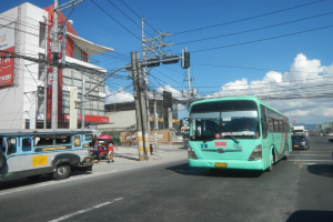 Ein grüner Bus fährt auf einer Straße mit Hochhäusern entlang, mit Passanten, die Regenschirme halten und Strommasten mit Drähten zu sehen sind.