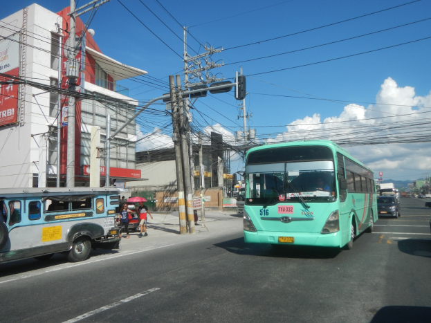 Ein grüner Bus fährt auf einer Straße mit Hochhäusern entlang, mit Passanten, die Regenschirme halten und Strommasten mit Drähten zu sehen sind.