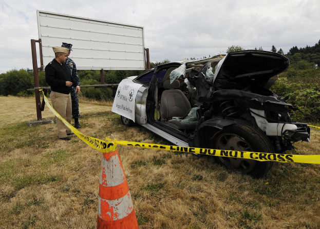 Ein Polizeibeamter in Uniform steht neben einem schwer beschädigten Auto am Straßenrand mit eingedrückter Haube und zersplitterten Fenstern, während ein Verkehrskegel und ein Absperrband an einem Pfosten in der Nähe gegen einen Hintergrund von Bäumen und einem bewölktem Himmel sind.