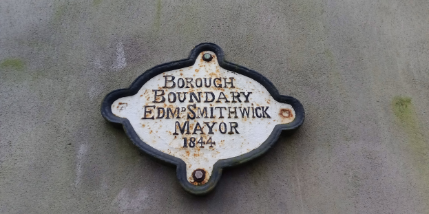Metallplatte an einer Gebäudeseite mit schwarzem Hintergrund und weißer fettgedruckter Schrift, die "Gemeindegrenze Edm Smithwick Bürgermeister" lautet.